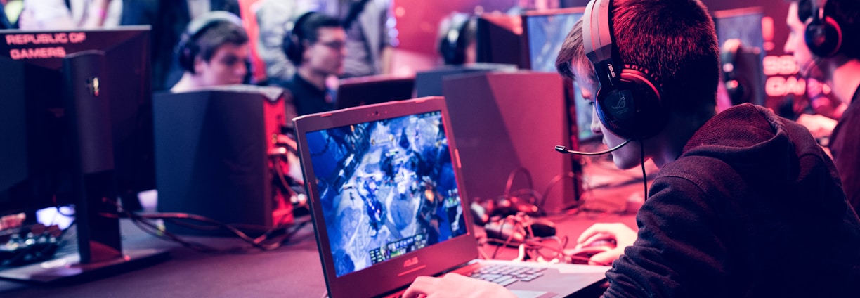 Several people are seated and focused on computer screens, wearing headsets, in an environment that appears to be a gaming tournament or esports event. The room is dimly lit with neon lighting, highlighting screens and creating an intense atmosphere.