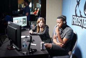 Two people are seated at a table with gaming monitors, wearing headsets. They appear to be participating in an esports broadcast or commentary. The environment is dimly lit with additional equipment and people visible in the background. A large blue banner with the word 'Frostbite' and a stylized graphic of a wolf is displayed.