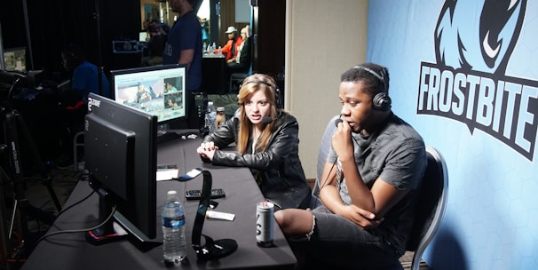 Two people are seated at a table with gaming monitors, wearing headsets. They appear to be participating in an esports broadcast or commentary. The environment is dimly lit with additional equipment and people visible in the background. A large blue banner with the word 'Frostbite' and a stylized graphic of a wolf is displayed.