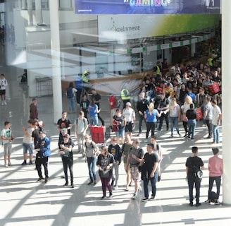 A large group of people gathered in a spacious, well-lit indoor area with glass walls. Many individuals are moving around, some in conversation and others holding bags. There is a large, visible queue in the background, organized by barriers, and several people are wearing gaming-related apparel. Signs with text hang from the ceiling.
