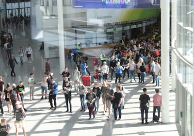 A large group of people gathered in a spacious, well-lit indoor area with glass walls. Many individuals are moving around, some in conversation and others holding bags. There is a large, visible queue in the background, organized by barriers, and several people are wearing gaming-related apparel. Signs with text hang from the ceiling.