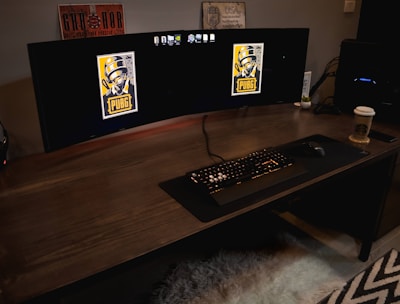 A gaming setup featuring a large curved monitor displaying PUBG logos on the screens. A keyboard and mouse are positioned on a large desk pad. A pair of headphones rests on the desk alongside a mobile phone. A drink in a Starbucks cup is placed to the right, while a desktop tower is seen in the background on the right side.
