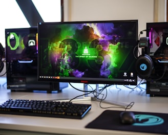 A gaming setup featuring a central monitor with a vibrant display showing 'The Spirit of Gaming'. On either side, there are gaming PCs with illuminated components and transparent cases. Headsets hang from the side of the monitor. The desk also has a black keyboard and mouse, and a branded mousepad.