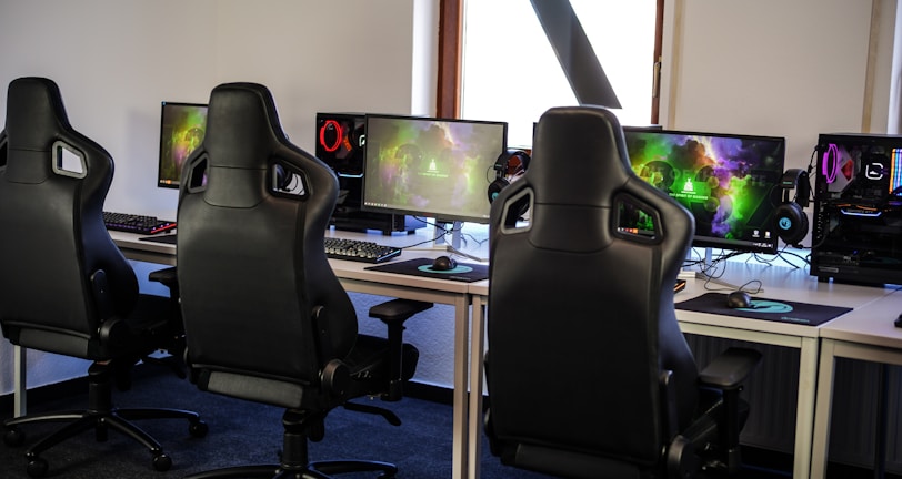 A row of gaming stations equipped with large monitors, gaming chairs, keyboards, and headphones. The setup is organized and ready for gaming, featuring a colorful screen display. The room has a modern and tech-savvy ambiance.