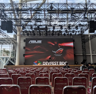 A stage setup with a large screen displaying logos for ASUS and Republic of Gamers. Rows of red chairs are arranged in front of the stage. The event is titled 'DEVFEST BDC', suggesting a tech or gaming conference. The venue appears to be indoors with a visible ceiling truss structure.