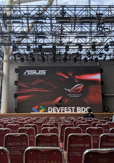 A stage setup with a large screen displaying logos for ASUS and Republic of Gamers. Rows of red chairs are arranged in front of the stage. The event is titled 'DEVFEST BDC', suggesting a tech or gaming conference. The venue appears to be indoors with a visible ceiling truss structure.