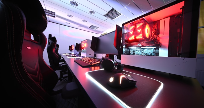 A row of high-end gaming computers on sleek desks, featuring RGB lighting and gaming accessories. The computers are housed in transparent cases with visible components, emitting a red glow. Comfortable gaming chairs with ergonomic design are positioned next to the desks, and the room's ceiling is modern with recessed lighting.