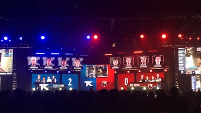 An esports event with a large screen displaying two teams, Team Kaliber and Rise Nation, competing against each other. The screen shows profiles of team members and scores, with Team Kaliber leading 2-0. The venue is dimly lit with blue and red lighting, and an audience can be seen watching.