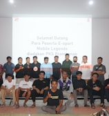 A group of people is posing for a photograph in a room with a white screen in the background that has text welcoming e-sport participants for a Mobile Legends event organized by PKS Prambanan. The group consists of 20 individuals standing and sitting, with some wearing casual attire and others wearing matching shirts that include logos.