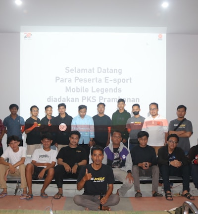 A group of people is posing for a photograph in a room with a white screen in the background that has text welcoming e-sport participants for a Mobile Legends event organized by PKS Prambanan. The group consists of 20 individuals standing and sitting, with some wearing casual attire and others wearing matching shirts that include logos.